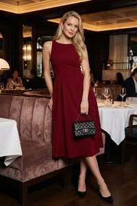 Mujer con vestido burdeos sin mangas sosteniendo un bolso negro acolchado, de pie junto a un reservado de restaurante con iluminación ambiental tenue.