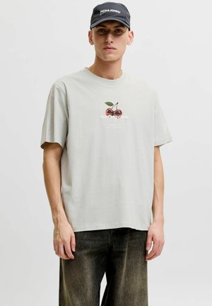 Tânăr purtând un tricou gri deschis cu design de cireșe și o șapcă neagră cu logo-ul "JACK & JONES", stă pe un fundal alb simplu.