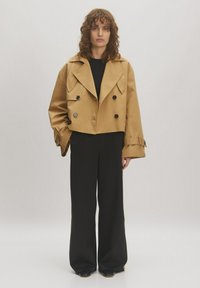 Ingeknipte, dubbelzijdige jas van beige stof met brede revers en zwarte knopen, gecombineerd met zwarte wijde broek en een eenvoudig zwart topje.