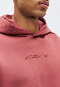 Νεαρό άτομο που φοράει μια ροζ φούτερ με κουκούλα με το κείμενο "J.LINDEBERG." ανάγλυφο στο στήθος, κομμένο κάτω από τη μύτη.