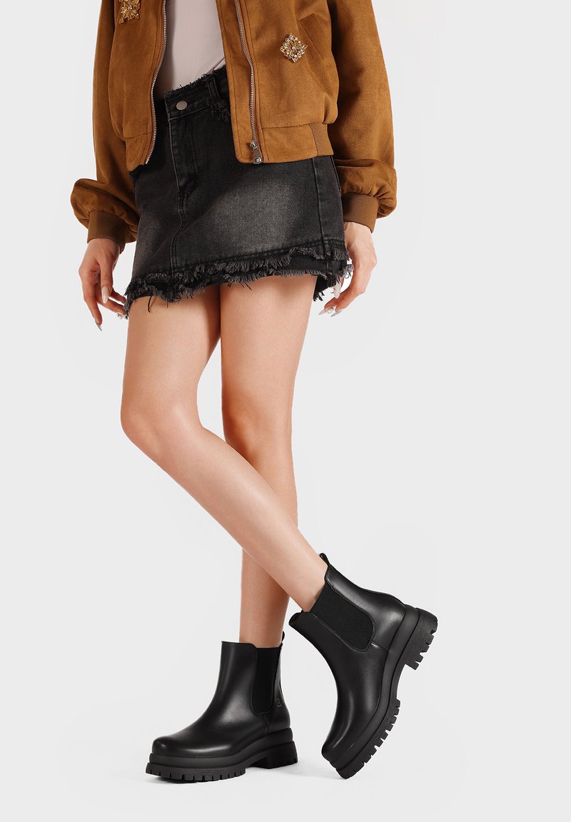 Donna che indossa stivali neri in pelle alla caviglia, mini gonna in denim nero strappata e giacca in suede marrone, in piedi su uno sfondo chiaro.