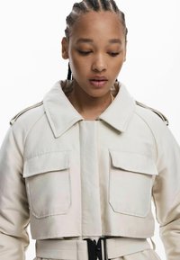 Veste crème cintrée avec de grandes poches sur la poitrine, large col et taille ceinturée. Tissu lisse avec une finition mate et un minimum de matériel.