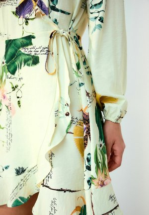 Közeli kép egy krémszínű, növényi mintás, kézzel írott szöveggel díszített wrap ruháról, amelyen megkötött derékszíj és ráncolt hosszú ujjú mandzsetta látható.