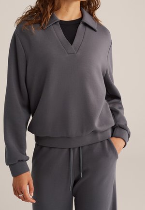 Femme portant un pull à manches longues gris foncé avec col et encolure en V, associé à un pantalon à cordon assorti, main dans la poche.