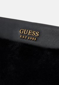 Prodotto in pelle nera con superficie texture, caratterizzato da un accento in pelliccia nera soffice e il logo dorato "GUESS EST 1981".