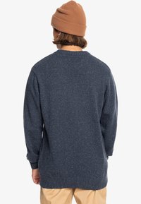 Granatowy sweter dzianinowy o teksturowanym wykończeniu, okrągłym dekolcie i długich rękawach. Noszony z brązową czapką dla uzyskania warstwowego wyglądu.