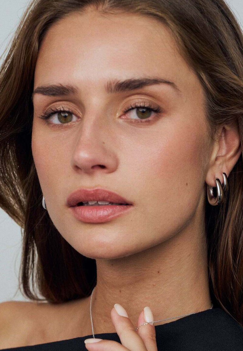 Silverörhängen i form av ringar, jämn hud, naturlig makeup, neutral läppfärg, lager av smycken med en delikat kedja, och mörka kläder.