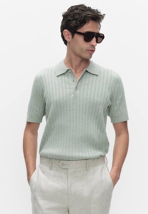 Man draagt een lichtgroen gebreid poloshirt met korte mouwen, beige broek met handen in de zakken en donkere zonnebril, kijkt naar rechts tegen een witte achtergrond.