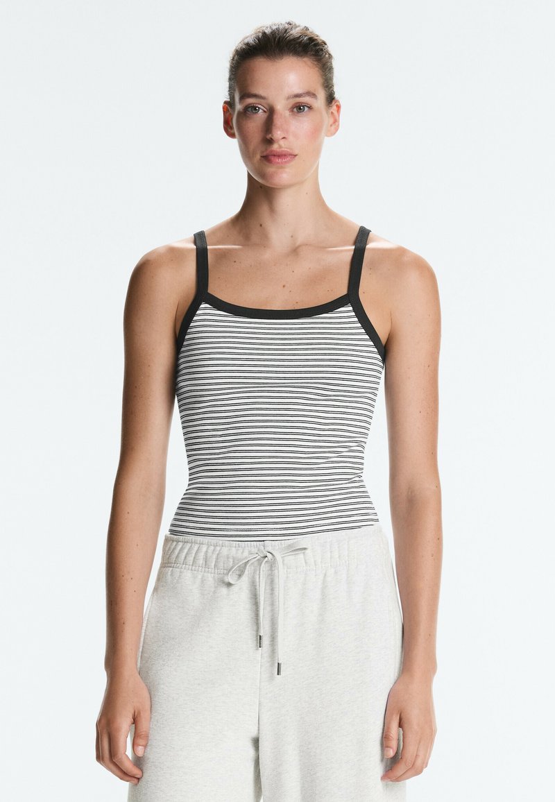 Femme aux cheveux attachés portant un haut sans manches rayé noir et blanc et un pantalon gris clair à cordon, debout devant un fond uni.