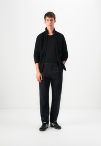Giovane uomo in piedi che indossa un maglione nero con zip, una t-shirt nera, pantaloni scuri e scarpe da ginnastica nere con strisce bianche su uno sfondo semplice.