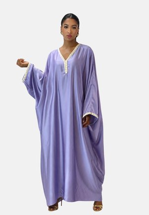 Kvinde iført en løs lilla satin kaftan med hvid kant på V-halsen og ærmerne, stående med den ene arm hævet, iført guldbelagte øreringe og hæle.