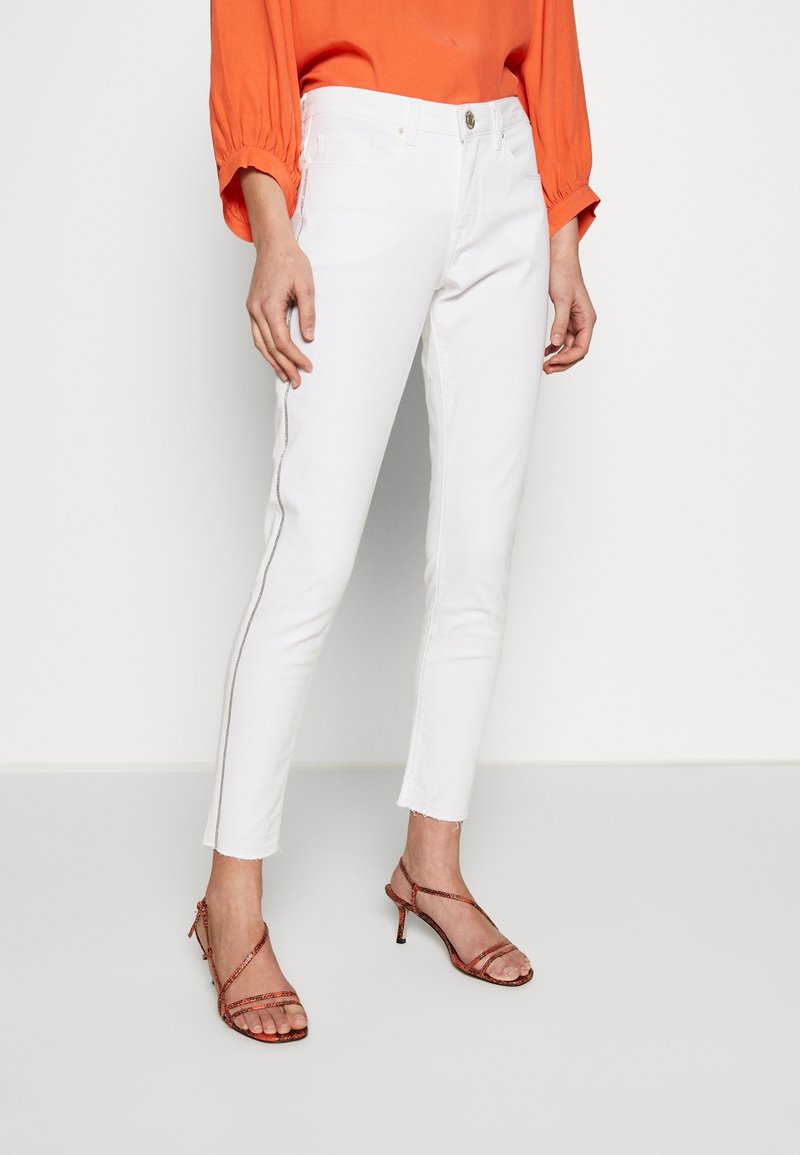 Jeans blancos ajustados con un dobladillo deshilachado, acentuados con una franja lateral brillante, combinados con una blusa naranja y sandalias de tacón con tiras.