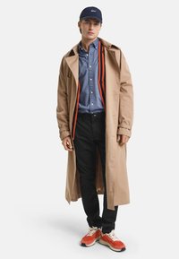 Beige trenchcoat over een blauwe shirt, zwarte broek en multicolor sneakers. Opvallende details zijn een marineblauwe pet en gestreepte binnenvoering.