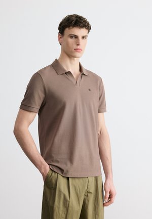 Jeune homme portant un polo marron à manches courtes et un pantalon ample vert olive, debout avec une main dans la poche, sur fond blanc.