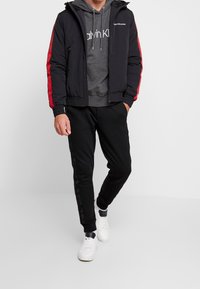 Svart bomberjacka med röda sidostripes, grå huva tröja, svarta sweatpants och vita sneakers. Har dragkedjeförslutning och logodetaljer.