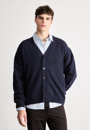 Navy knitted cardigan with a deep V-neck and five buttons, worn over a light blue striped shirt. Soft texture and relaxed fit.