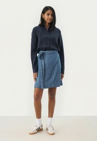 Chemise boutonnière navy, jupe en denim bleu clair enroulée et baskets crème avec rayures noires. La tenue présente un design décontracté et superposé.