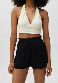 Top corto de marca con tirantes en tela color blanco roto y diseño de escote en V. Combinado con pantalones cortos negros texturizados con cintura elástica y cordón.