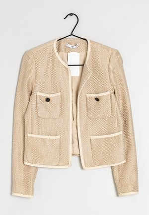Veste beige texturée pour femme avec manches longues, quatre poches avant et boutons noirs, suspendue sur un cintre noir sur fond blanc.
