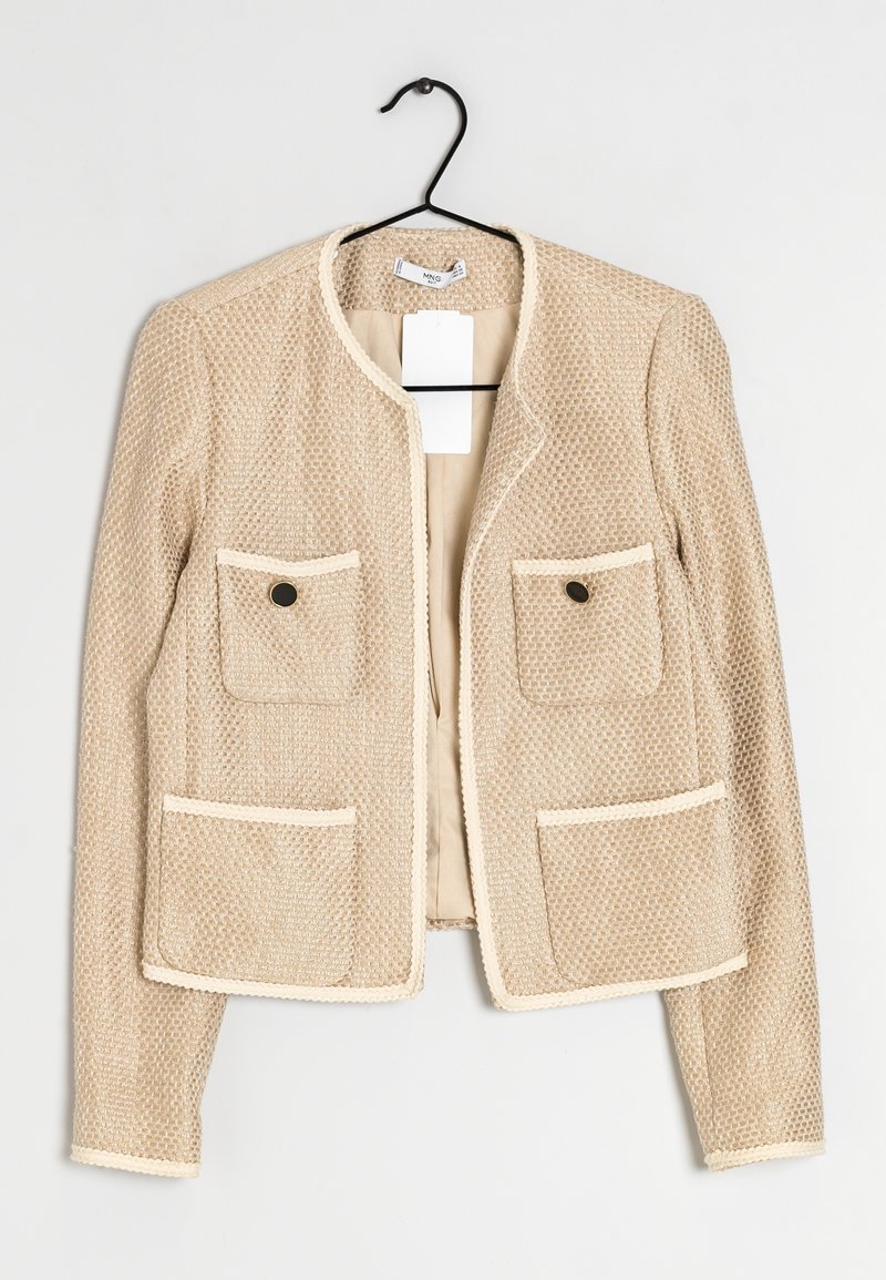 Veste beige texturée pour femme avec manches longues, quatre poches avant et boutons noirs, suspendue sur un cintre noir sur fond blanc.