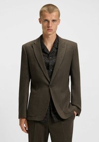 Braun strukturierter Blazer mit zwei Vorderaschen, kombiniert mit einem dunklen Hemd mit einem sich wiederholenden Logo-Muster. Taillierte Passform, Ein-Knopf-Verschluss.