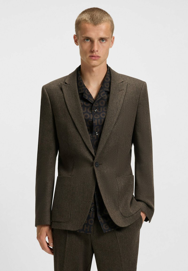 Blazer marrone texturizzato con due tasche frontali, abbinato a una camicia scura con un motivo a logo ripetitivo. Vestibilità su misura, chiusura a un bottone.