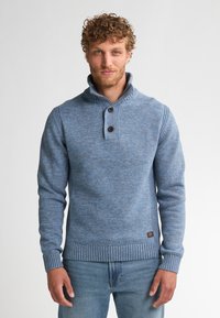 Hombre con pelo rizado que lleva un suéter de punto azul con botones y jeans azules, de pie contra un fondo blanco liso.