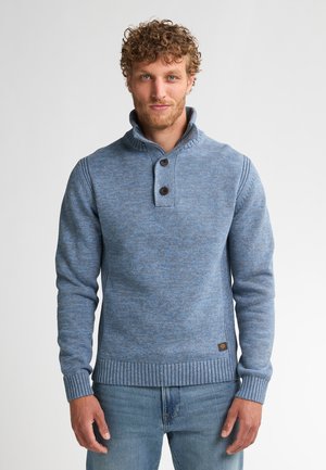 Hombre con pelo rizado que lleva un suéter de punto azul con botones y jeans azules, de pie contra un fondo blanco liso.
