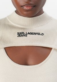 Šviesiai bevardis ribotas viršus su aukštu apykakle ir dideliu išpjovimu. Ant viršutinės krūtinės dalies yra juodas siuvinėtasis 'KARL LAGERFELD JEANS' logotipas.