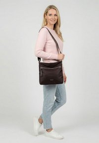 Lächelnde Frau im rosa Pullover, hellen Jeans und weißen Sneakers, mit einer dunkelbraunen Umhängetasche mit Reißverschlusstasche vorne, seitlich stehend.