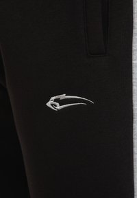 Musta verkkohousut, joissa on sileä kangas, urheilullinen istuvuus, sivutaskut ja valkoinen brodeerattu logo reidessä.