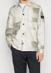 Camisa de manga larga, de color claro, con un patrón de cuadros en tonos variados de verde, que presenta dos bolsillos frontales y detalles de botones negros.