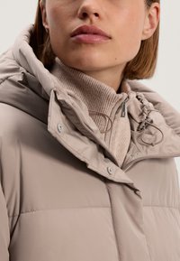 Svijetlo bež puffer jakna s prilagodljivom kapuljačom, dugmadi na pritisak i detaljima s utezima. Nosi se preko teksturirane bež rolke.