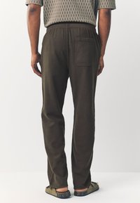 Green elastic-waist pants with a single back pocket, featuring a textured fabric and a relaxed fit, paired with flat-soled shoes.
