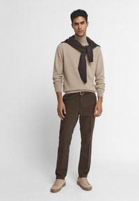 Beige stickad tröja, brun scarf hängande över axlarna, bruna byxor och ljusbeiga sneakers, mot en neutral bakgrund.