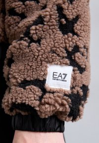 Brown and black textured fleece sleeve with a visible elastic cuff and a white label featuring "EA7 Emporio Armani."