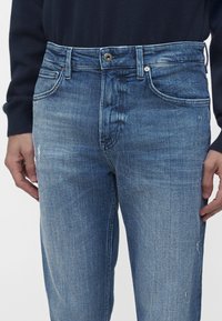 Homme portant un jean en denim bleu délavé et un haut à manches longues foncé, les mains reposant sur les hanches.