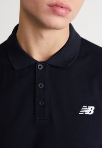 Polo de color azul marino oscuro, hecho de un tejido texturizado, con un cuello clásico, tres botones negros y un logo "NB" blanco en el lado izquierdo del pecho.