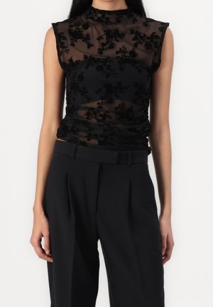 Vrouw draagt een mouwloos zwart doorschijnend topje met bloemenpatronen, gecombineerd met hooggetailleerde zwarte op maat gemaakte broek met een riem.