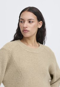 Maglione lavorato a maglia beige con collo rotondo, coste testurizzate e dettagli a pannello diagonale sulle spalle. Tessuto morbido e leggero.