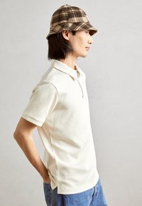 Short-sleeve cream polo shirt with textured geometric pattern and half-zip collar, paired with blue denim jeans and a plaid bucket hat.