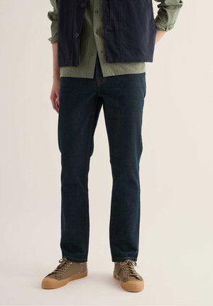 Person trägt dunkelblaue Jeans, olivgrünes Hemd mit Knöpfen, marineblaue Weste und olivgrüne Sneakers mit Gummisohlen, steht vor einem neutralen Hintergrund.