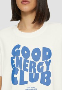 Witte t-shirt met korte mouwen, met grote blauwe grafische tekst die "GOOD ENERGY CLUB" leest en een kleinere slogan eronder.