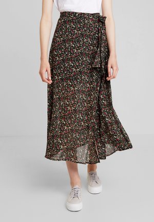 Femme portant une jupe portefeuille midi noire à motifs floraux avec de petites fleurs rouges et blanches et des baskets beige clair sur un fond uni.