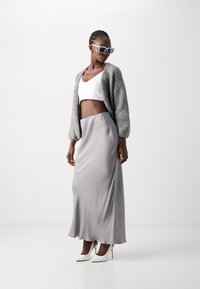 Cardigan gri pufo, bluză albă scurtă și o fustă maxi din satin gri fluidă, asociate cu tocuri albe cu vârf ascuțit. Ochelari de soare moderni completează ținuta.
