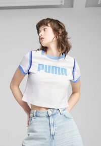 Bílý zkrácený tričko s modrými žebrovanými manžetami a límcem, s velkým modrým logem "PUMA". Kombinováno se světle modrými džínovými šortkami s vysokým pasem.