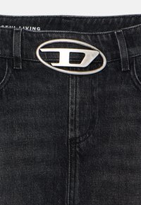 Un jean en denim noir présente une boucle ovale en argent brillant avec un logo, des poches avant et un design classique à cinq poches.