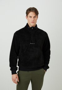 Sort fleece pullover med høj krave, kvart lynlås og elastiske manchetter. Har et lille logo på brystet og en blød, tekstureret overflade.