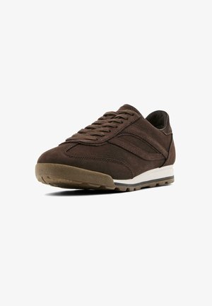 Scarpa atletica in suede marrone con superficie testurizzata, suola in gomma e intersuola bianca a contrasto. Presenta un classico design con lacci e dettagli cuciti.