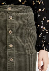 Jupe en velours côtelé vert olive avec des nervures verticales, des boutons sur le devant et une poche latérale, associée à un blouse noire à motif floral.